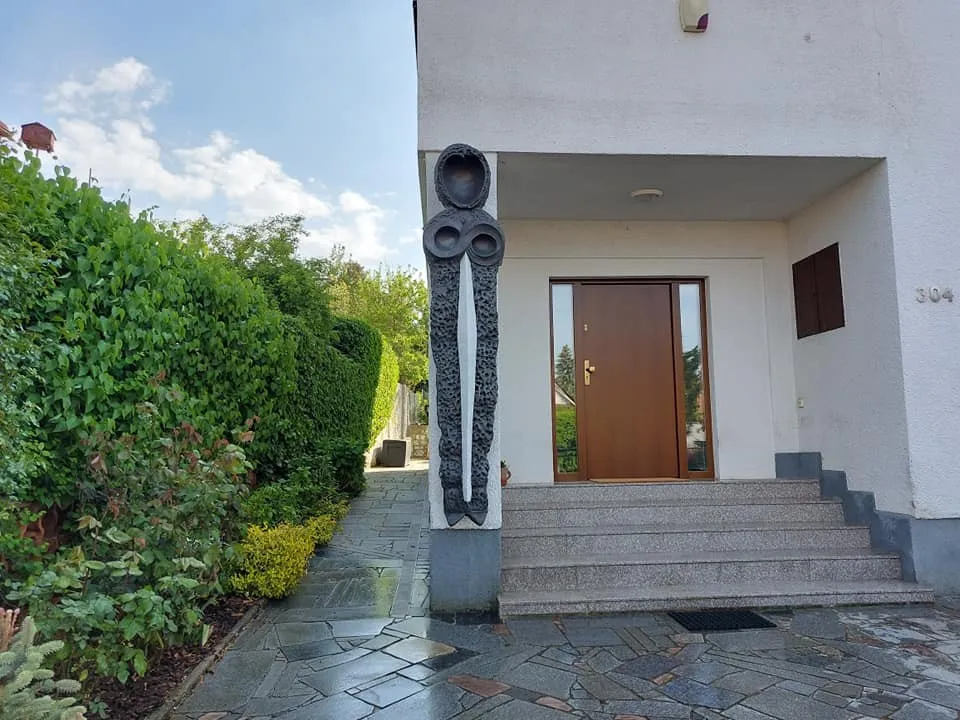 Vysoká tmavá plastika s kruhovou hlavou u vchodu do rodinného domu