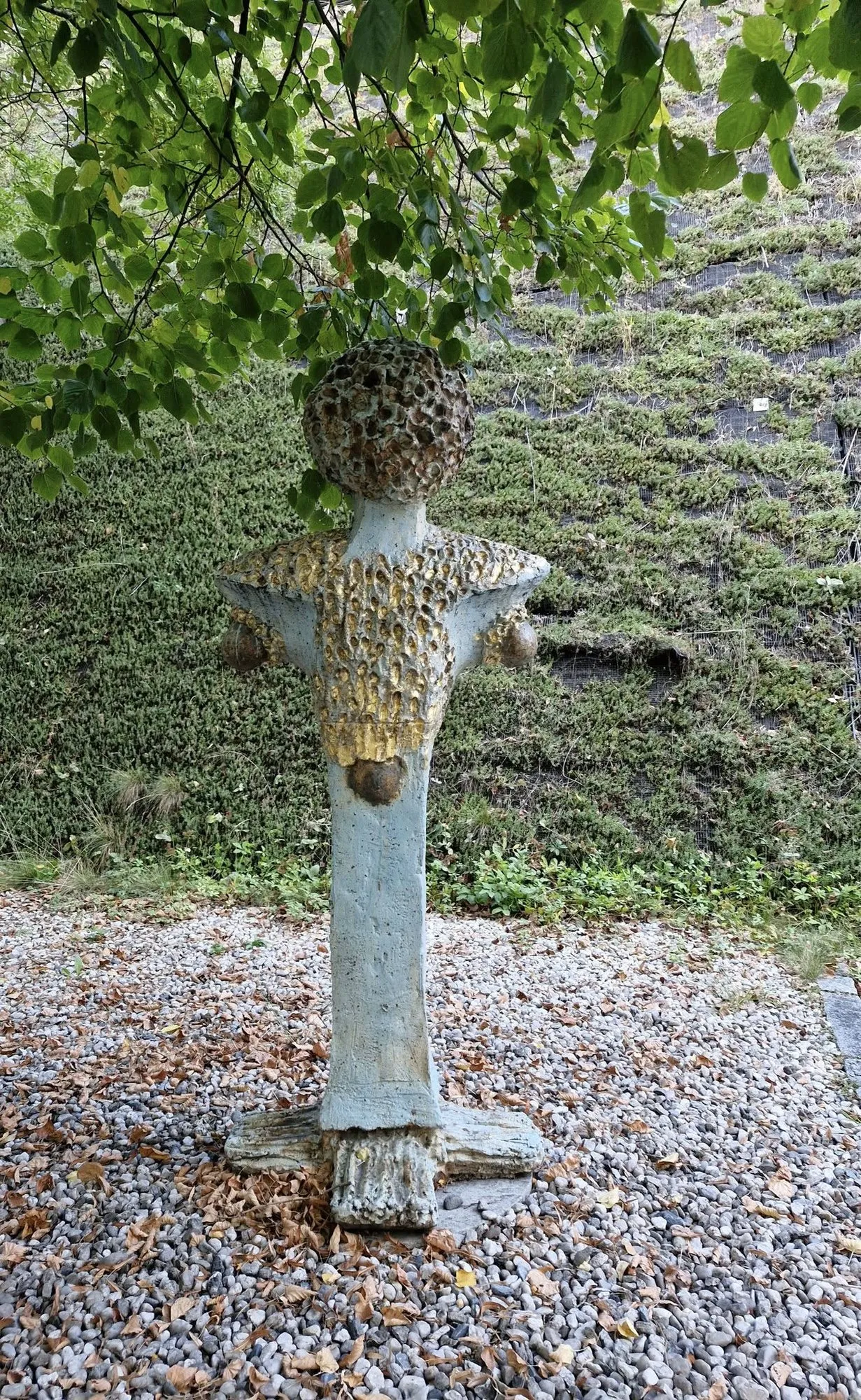 Modrá figura se strukturovanou hlavou na štěrkové zahradě pod stromem